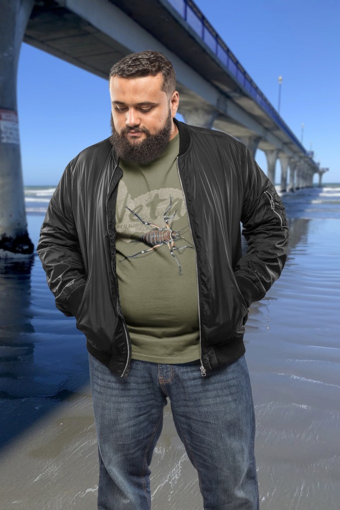 A man wears his army green giant weta t-shirt under his bomber jacket beside New Brighton Pier.