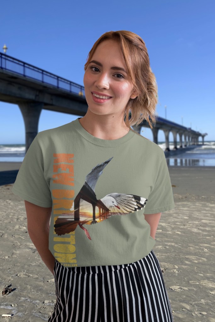A happy young woman wearing a light olive Red-billed gull, New Brighton, New Zealand t-shirt on New Brighton beach beside the New Brighton pier.