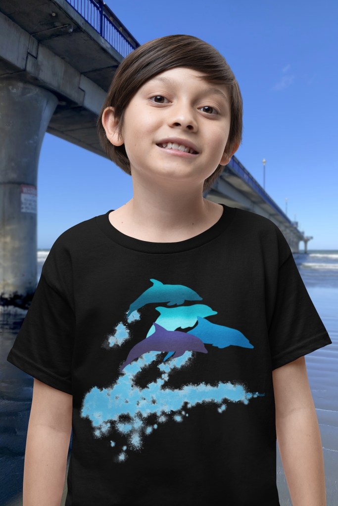 A smiling boy wearing a black t-shirt with a lively Surface Active “dolphins leaping” print standing beside New Brighton pier.