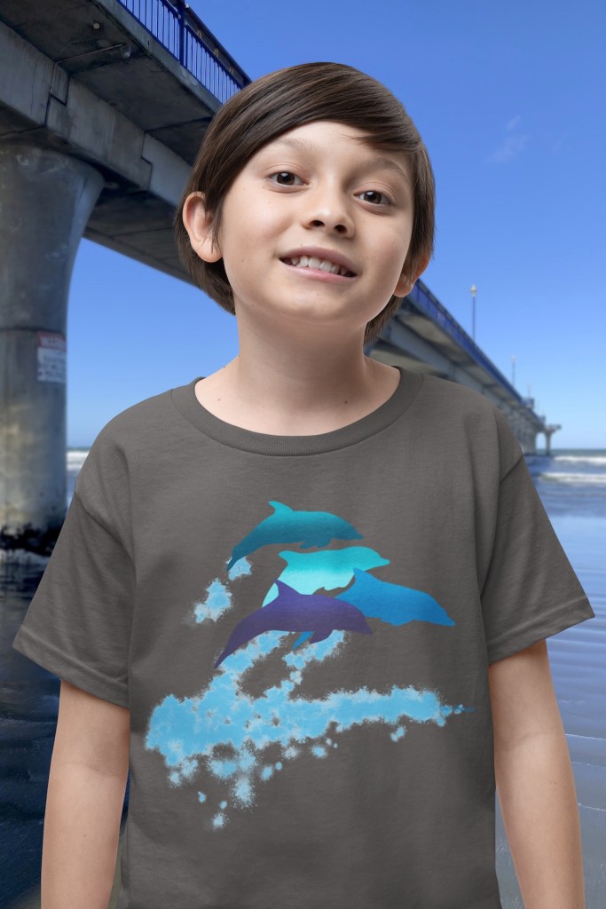 A smiling boy wearing a dark grey t-shirt with a lively Surface Active “dolphins leaping” print standing beside New Brighton pier.