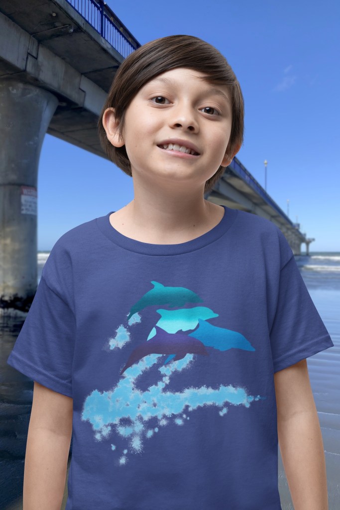 A smiling boy wearing a deep royal blue t-shirt with a lively Surface Active “dolphins leaping” print standing beside New Brighton pier.