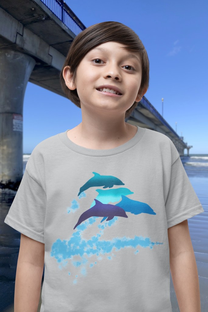 A smiling boy wearing a grey marle t-shirt with a lively Surface Active “dolphins leaping” print standing beside New Brighton pier.