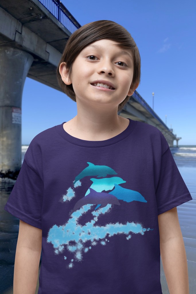 A smiling boy wearing a navy blue t-shirt with a lively Surface Active “dolphins leaping” print standing beside New Brighton pier.