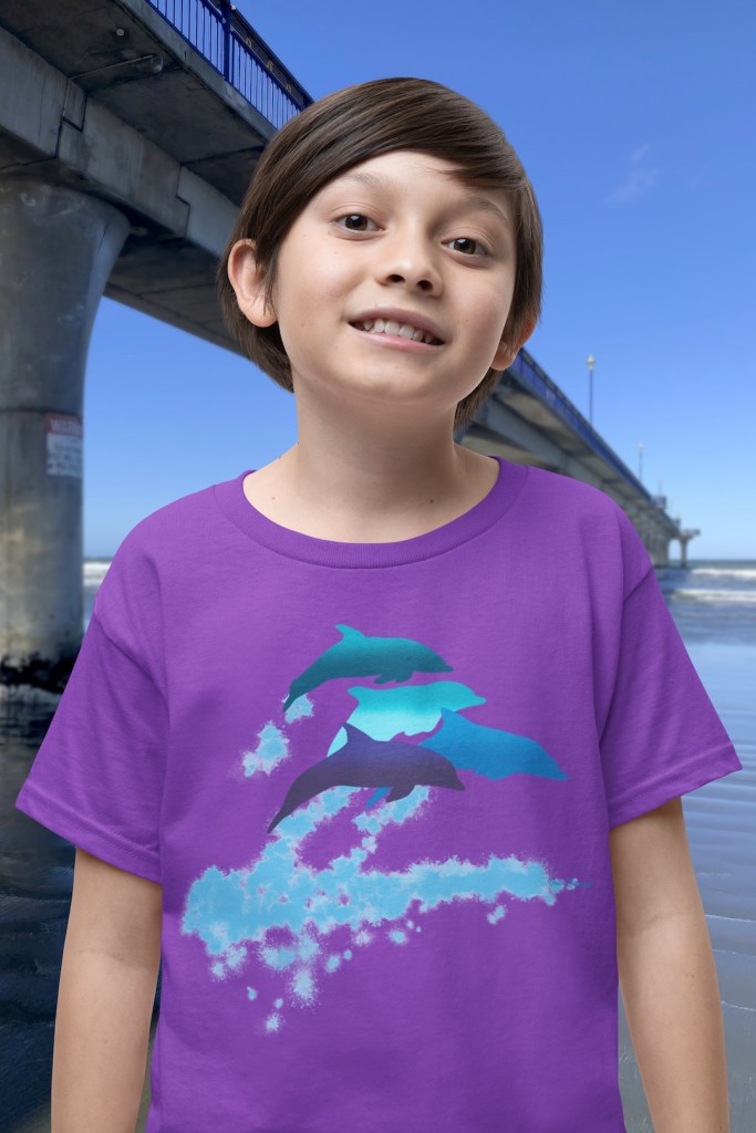 A smiling boy wearing a purple t-shirt with a lively Surface Active “dolphins leaping” print standing beside New Brighton pier.