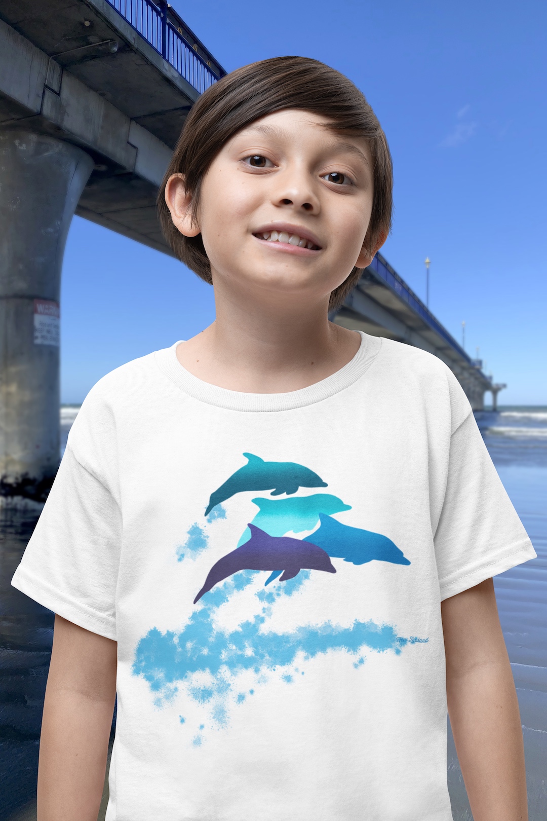 A smiling boy wearing a white t-shirt with a lively Surface Active “dolphins leaping” print standing beside New Brighton pier.