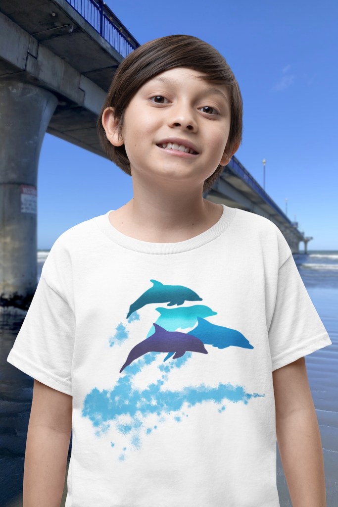 A smiling boy wearing a white t-shirt with a lively Surface Active “dolphins leaping” print standing beside New Brighton pier.
