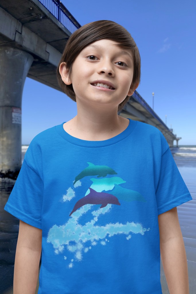 A smiling boy wearing an aqua blue t-shirt with a lively Surface Active “dolphins leaping” print standing beside New Brighton pier.