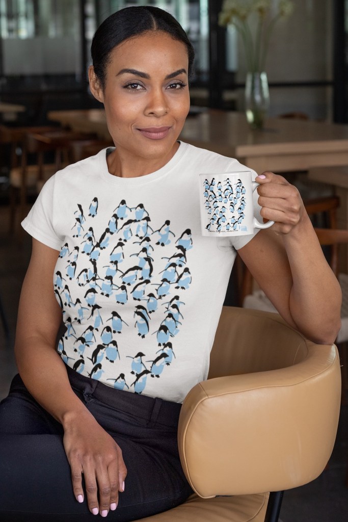 A playful t-shirt featuring the behaviour of a group of Adelie penguins in motion. The same design is repeated on a coffee mug that the model is in a cafe holding.