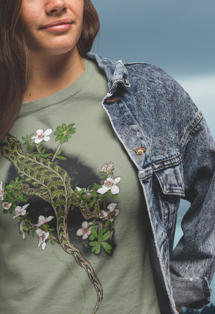 :Close-up of a woman wearing a Harlequin Gecko t-shirt under a denim jacket, highlighting the detailed print of the gecko's camouflage pattern against a backdrop of flowering manuka.