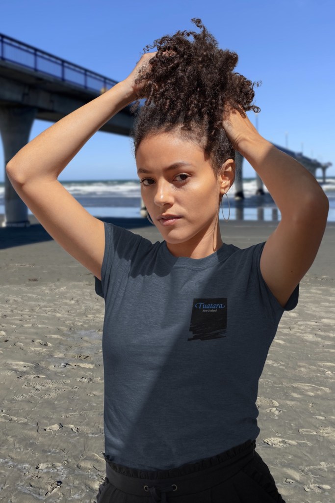 A young woman modelling the chest print on the front of a Surface Active Tuatara, New Zealand, t-shirt on New Brighton beach beside New Brighton Pier.
