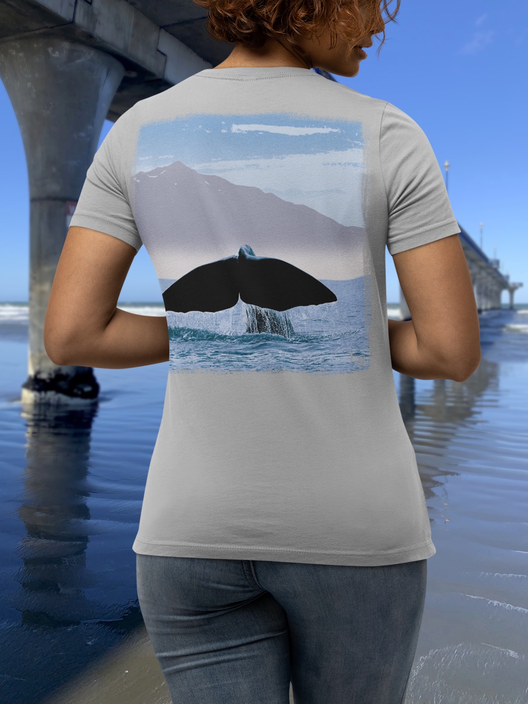 Back view of a curly-haired woman modelling the large art of a Surface Active Kaikoura New Zealand, home of the Mighty Sperm Whale t-shirt beside the New Brighton pier.