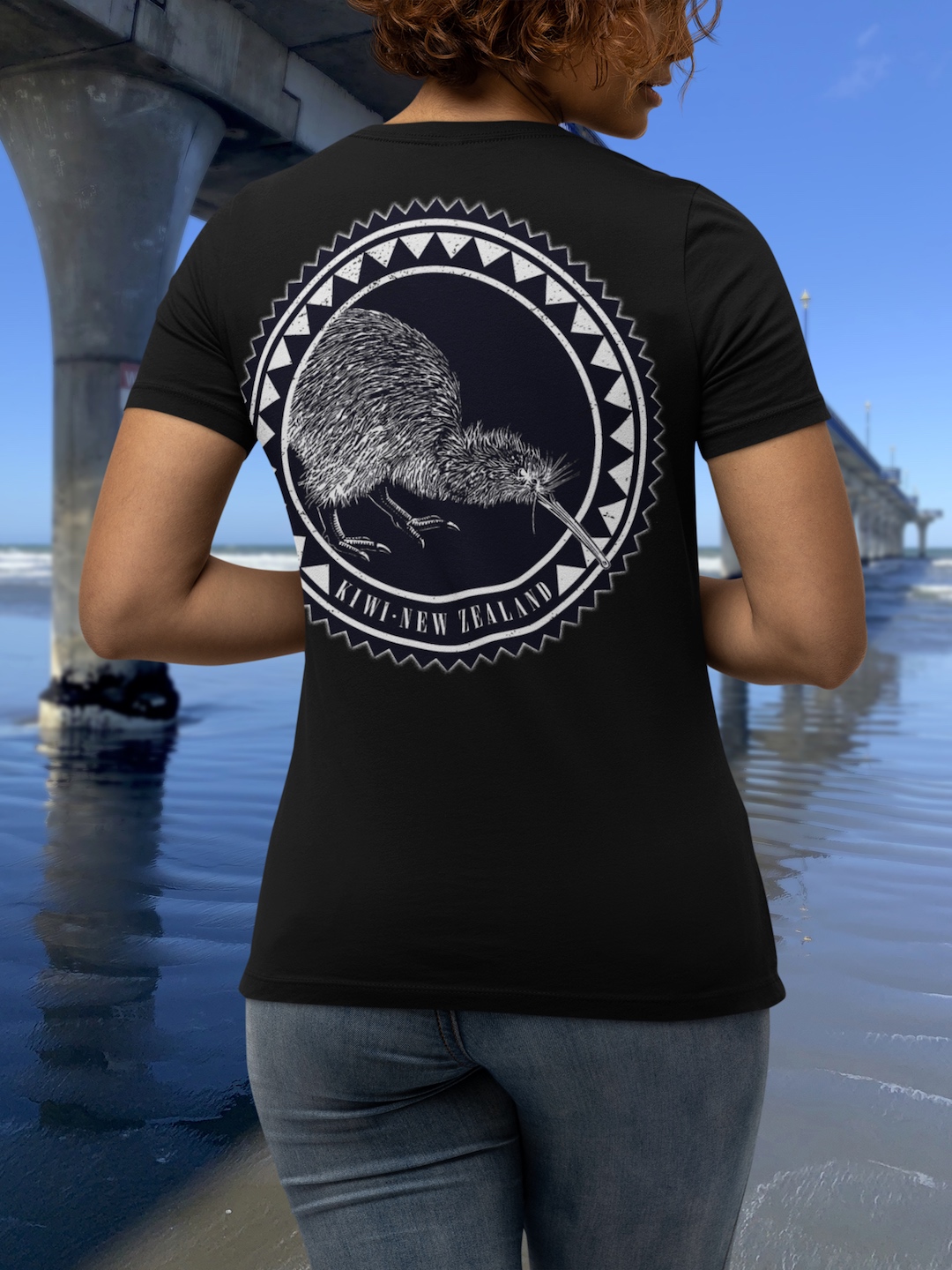 Back view of a woman in a black t-shirt with a large, two-colour kiwi roundel print, standing beside New Brighton Pier.