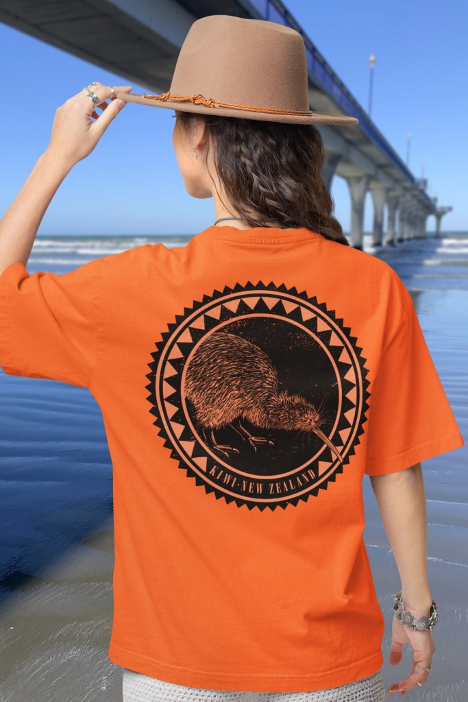 ack view of a woman in a bright orange t-shirt with a Kiwi Roundel print, standing by New Brighton Pier.