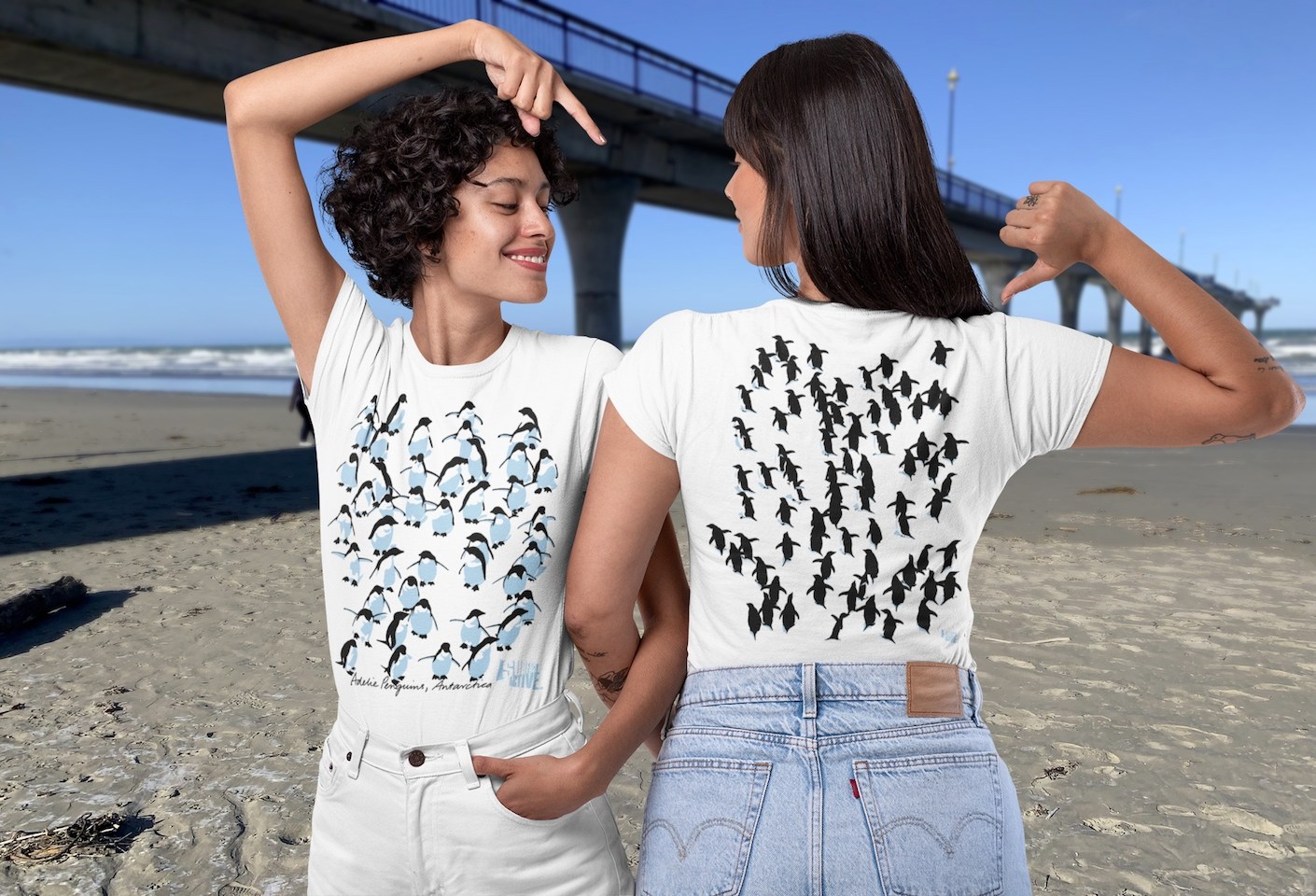 Two smiling women modelling both sides of the Surface Active Adelie penguins, Antarctica t-shirt on New Brighton beach in New Zealand, alongside the New Brighton pier.