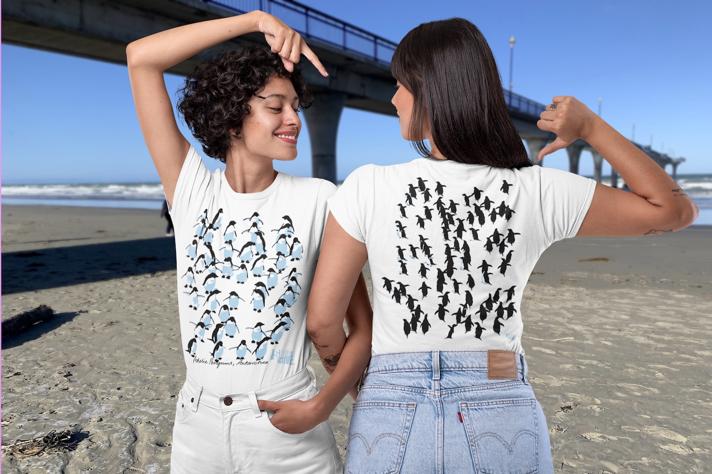 Two smiling women modelling both sides of the Surface Active Adelie penguins, Antarctica t-shirt on New Brighton beach in New Zealand, alongside the New Brighton pier.