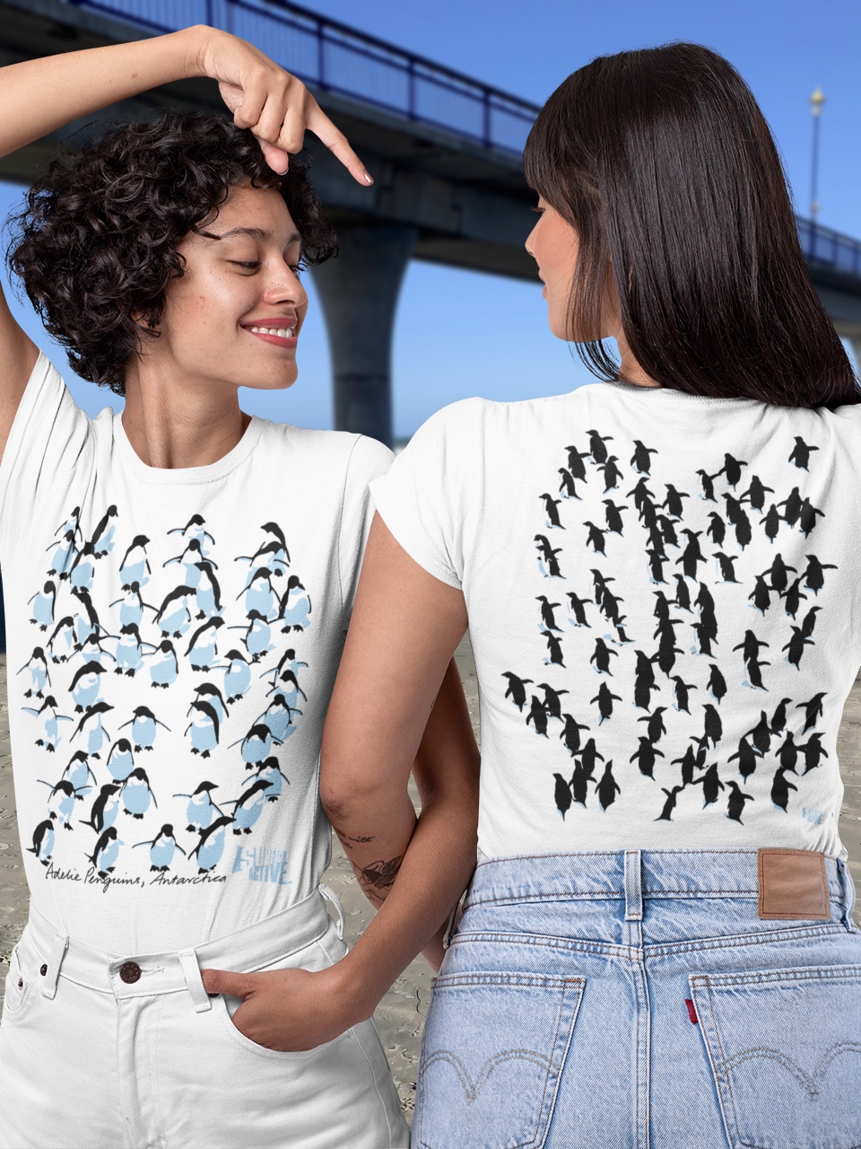 Two women wearing a two-sided Adelie Penguin t-shirt, displaying both the front and back designs, beside New Brighton Pier in Christchurch, New Zealand.