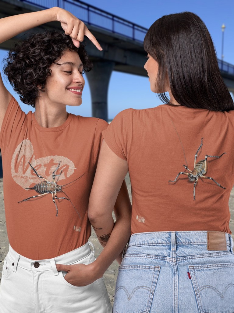 Two smiling women modelling both sides of the Surface Active Weta, New Zealand t-shirt on New Brighton beach alongside the New Brighton pier.