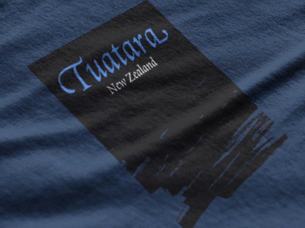 Extreme close-up of the chest badge print on a Surface Active ‘Tuatara, Stephens Island, New Zealand’ multicolour design on a navy blue t-shirt.