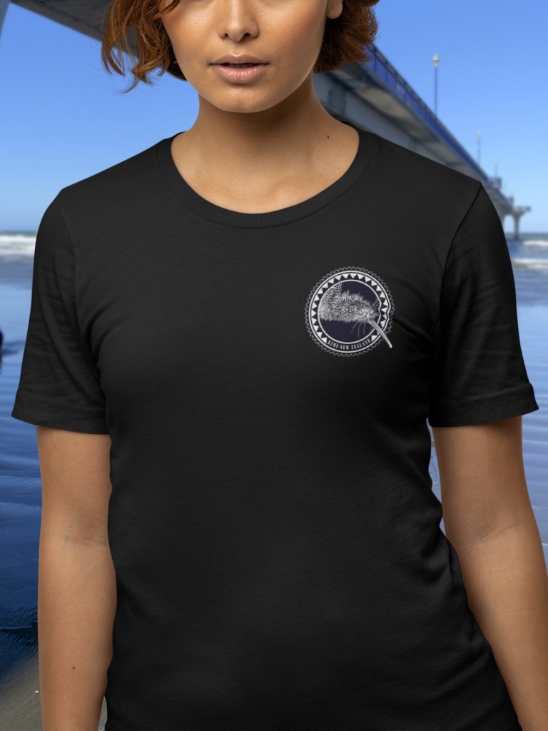 Close-up view showing the chest badge print on the Surface Active Kiwi, New Zealand t-shirt worn by a woman with curly hair standing beside the New Brighton pier.