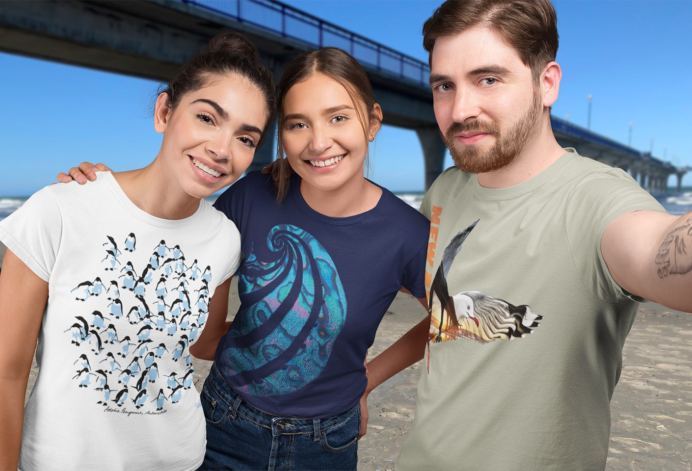 Three friends in Surface Active t-shirts with designs of Adelie penguins, Paua shell, and a red-billed gull at dawn, posing for a selfie at New Brighton Pier.