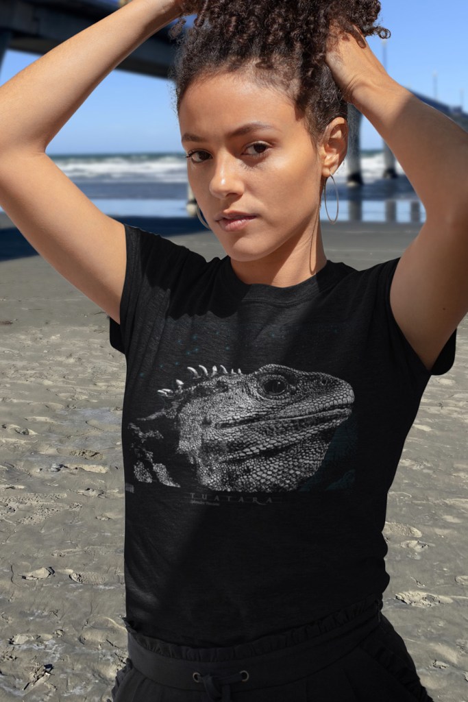 A curly haired young woman wearing her Surface Active Tuatara t-shirt on the beach beside New Brighton pier.