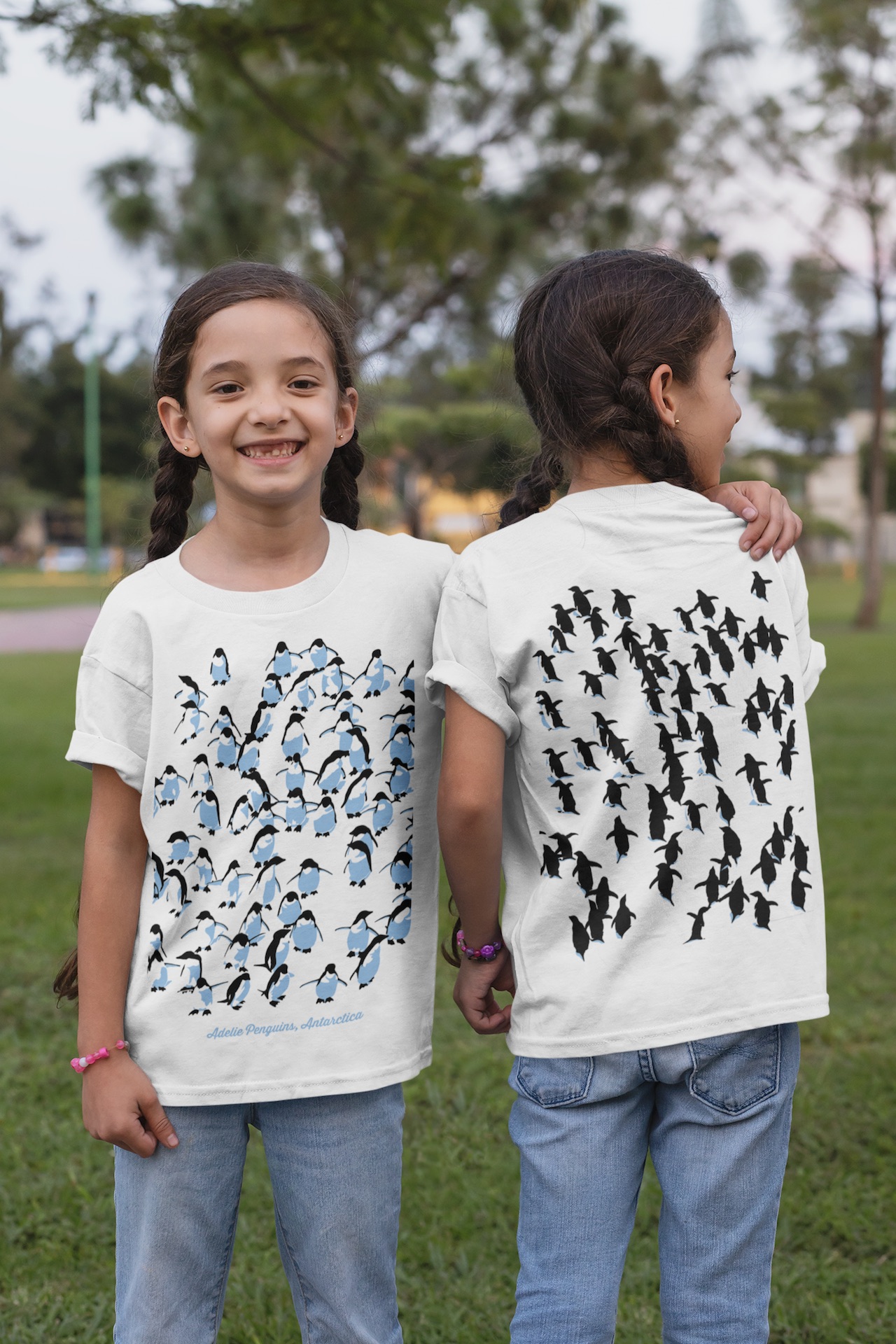 Twin girls in a park modeling a white Adelie Penguin kids’ t-shirt, showing front and back views with a three-colour penguin and leopard seal design.