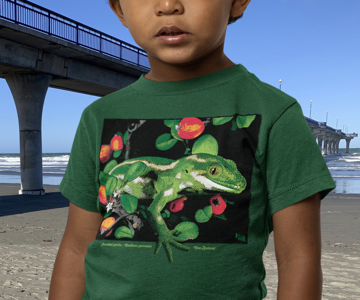 A little boy wearing a Surface Active Jewelled gecko, forest green t-shirt, on the beach, beside New Brighton pier.