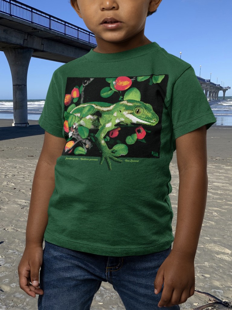 A little kid wearing a Surface Active Jewelled gecko, forest green t-shirt, on the beach, beside New Brighton pier.