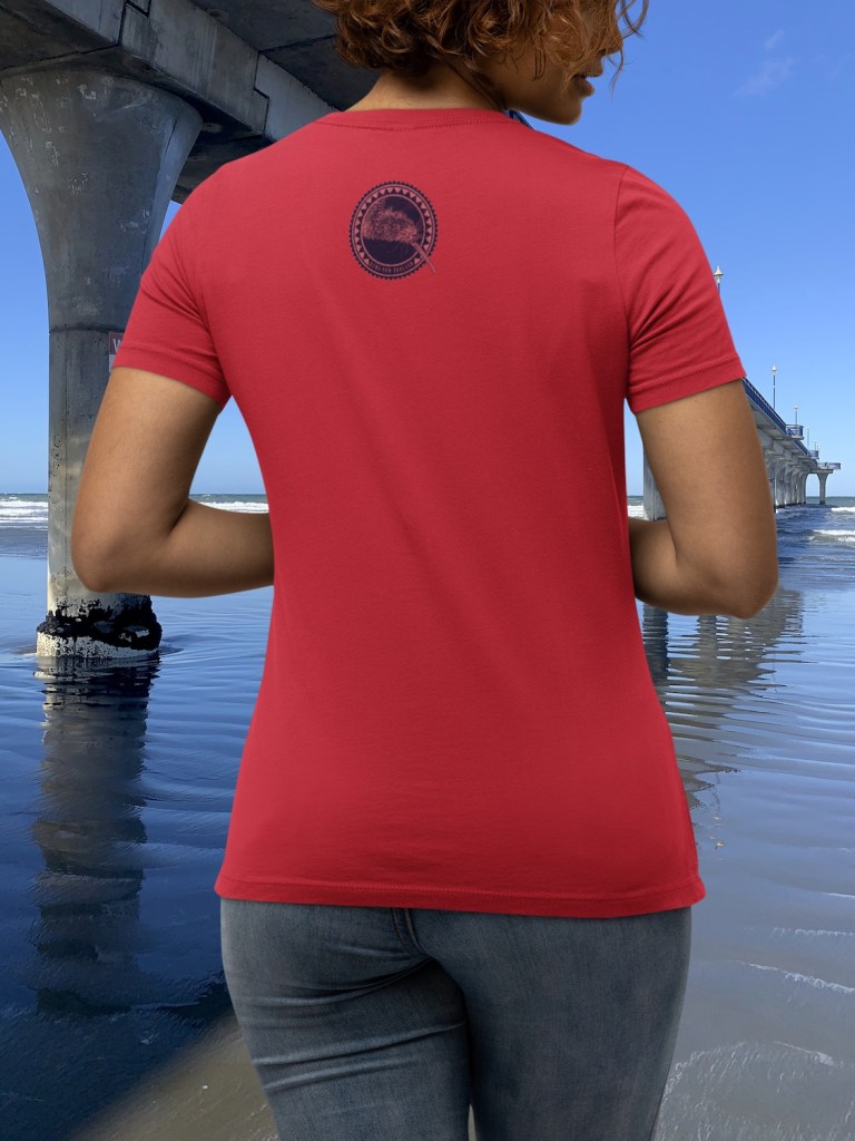 A woman showing the back of her red Surface Active iconic Kiwi t-shirt beside New Brighton Pier. The big art of a Kiwi roundel is printed on the shirt front and on the back there is a small round Kiwi portrait printed between the shoulders. This variant of the design is designed for printing on light, bright and pastel colours.