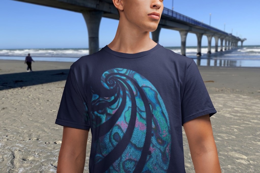 A young man wearing a navy blue Surface Active Paua t-shirt beside New Brighton Pier.