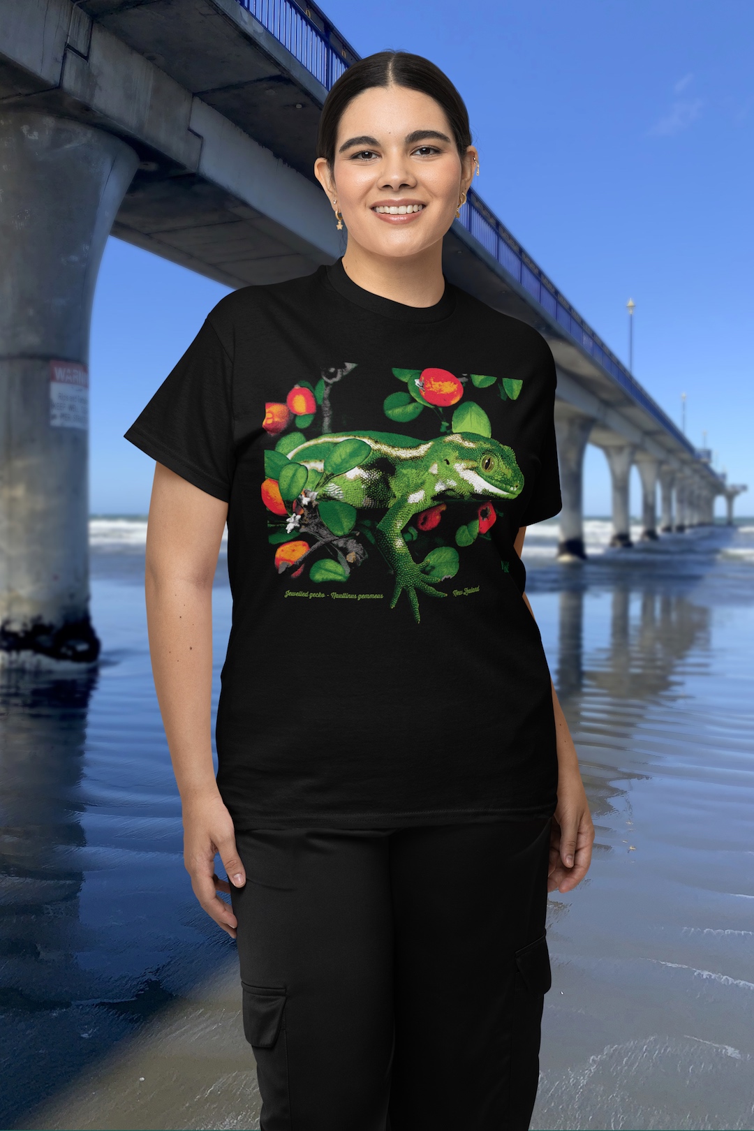 A woman wearing a black Surface Active jewelled gecko t-shirt beside New Brighton Pier.