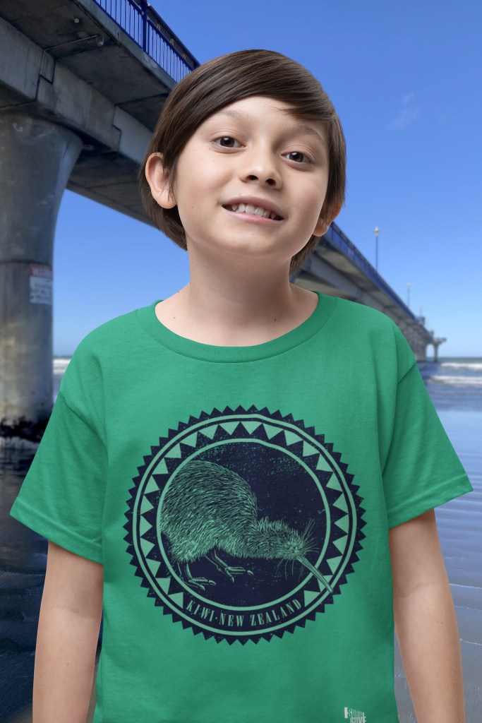 A smiling boy wearing a Kelly green Surface Active Iconic Kiwi t-shirt beside New Brighton Pier.