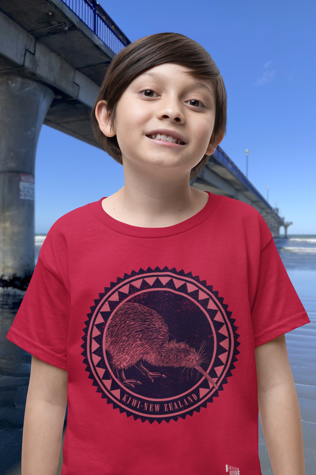 A happy boy in a red t-shirt with a large Kiwi Roundel design on the front, standing by New Brighton Pier.
