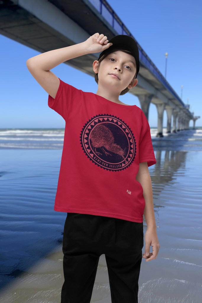 A stylish boy in a baseball cap wearing a red Surface Active Iconic Kiwi t-shirt beside New Brighton Pier.