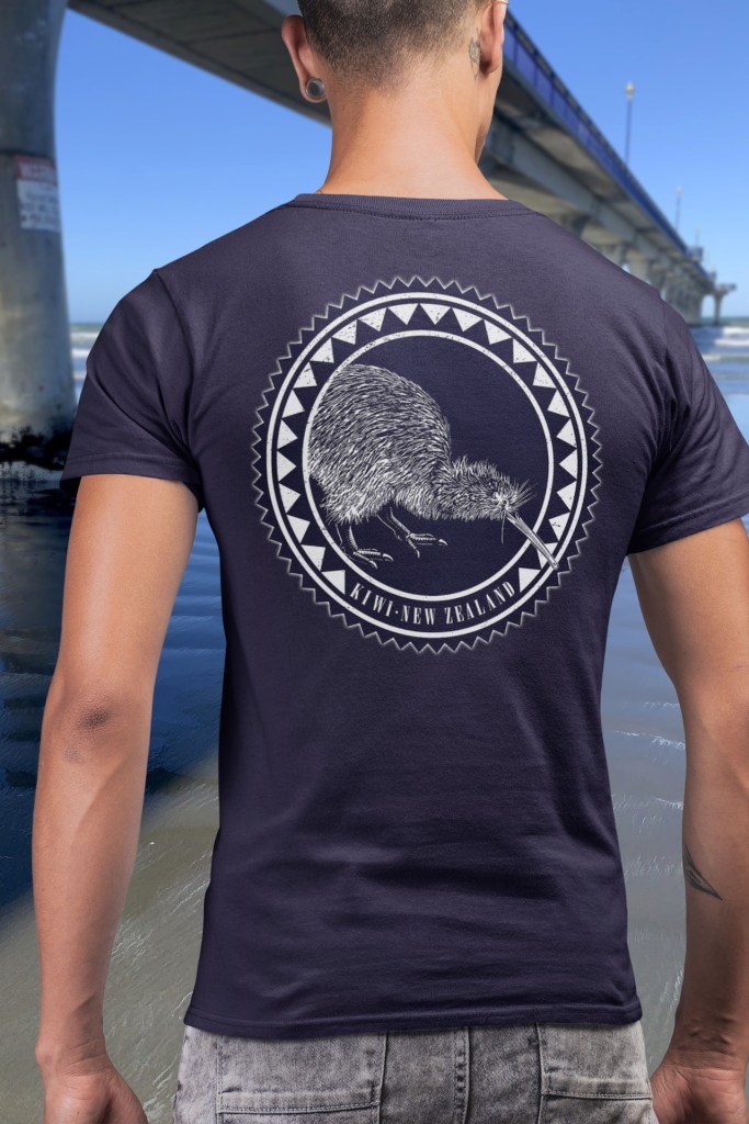 A man wearing a navy blue t-shirt with a large, two-colour kiwi roundel print on the back, standing beside New Brighton Pier.