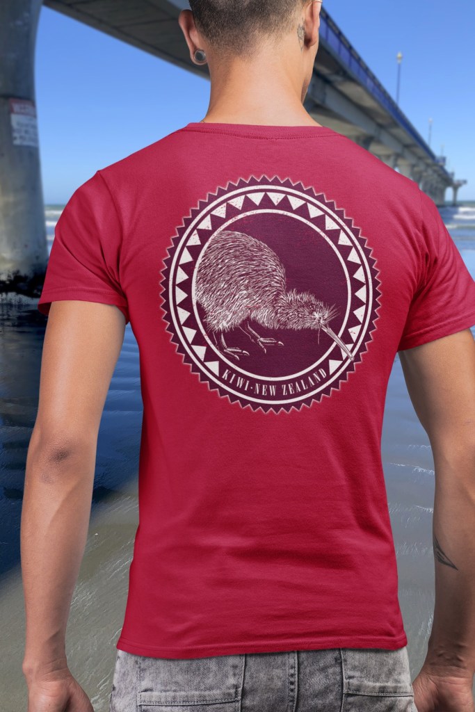 A man seen from the back wearing a red t-shirt with a large Kiwi roundel print, standing beside New Brighton Pier.