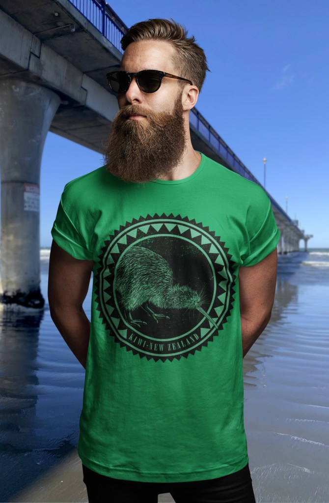 A bearded man in a kelly green t-shirt with a large, dark kiwi roundel design on the front, standing by New Brighton Pier.