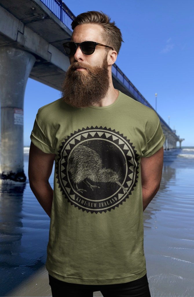 A bearded man in an army green t-shirt featuring a large, dark print of the Surface Active Kiwi Roundel design, standing beside New Brighton Pier.