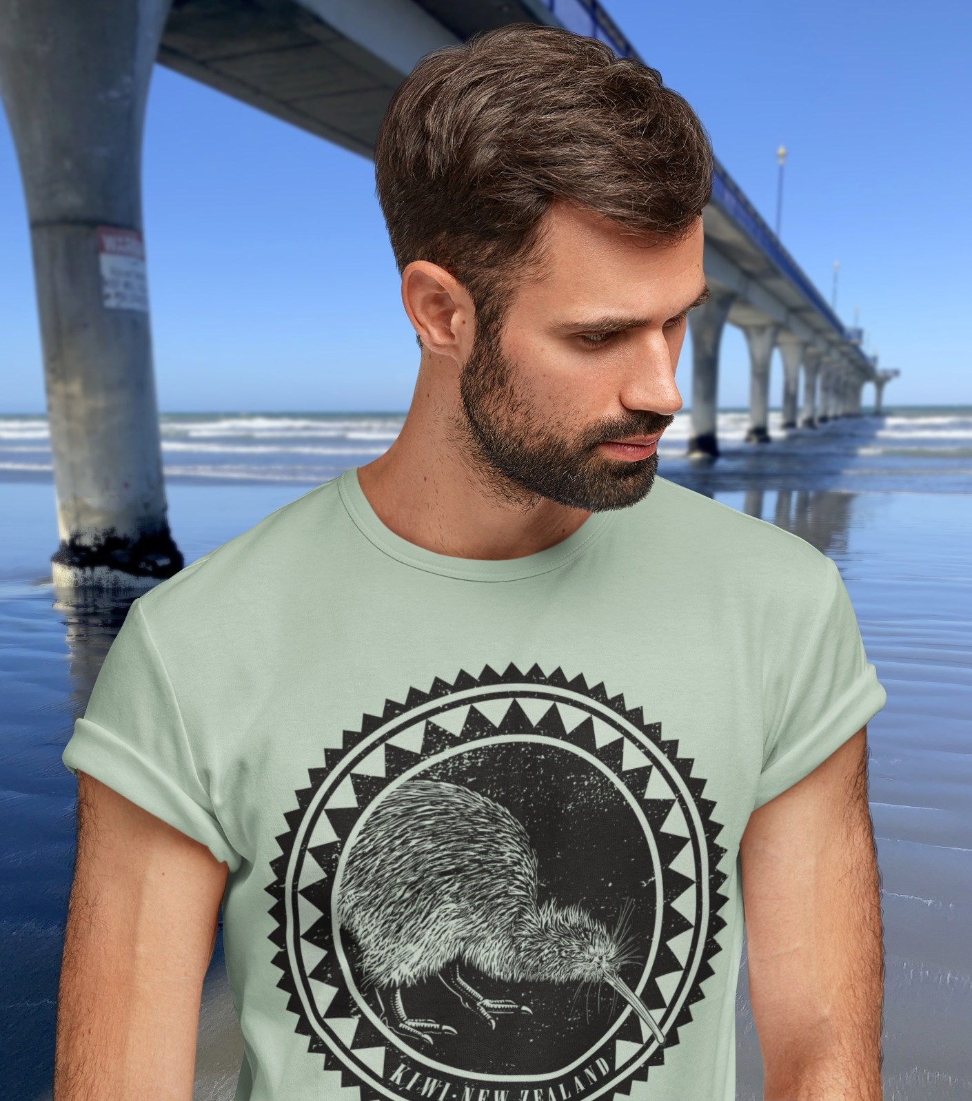 A sage green Surface Active iconic Kiwi roundel design t-shirt modelled by a bearded man beside New Brighton pier.