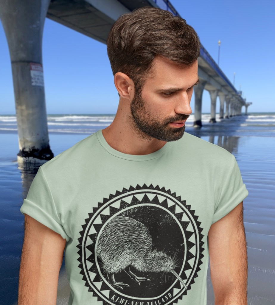 A sage green Surface Active iconic Kiwi roundel design t-shirt modelled by a bearded man beside New Brighton pier.