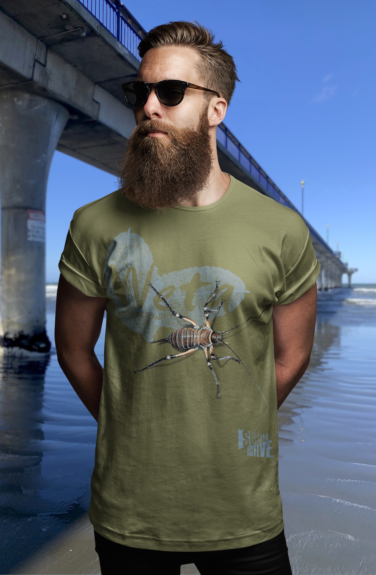 A hipster guy wearing an army green t-shirt with a large, pixel-art Bluff weta design stands with New Brighton Pier in the background.