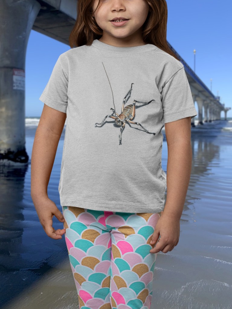 A young girl in a grey marle t-shirt with an all-over, two-sided print of two weta insects stands smiling next to New Brighton Pier, with the sea and sky in the background.
