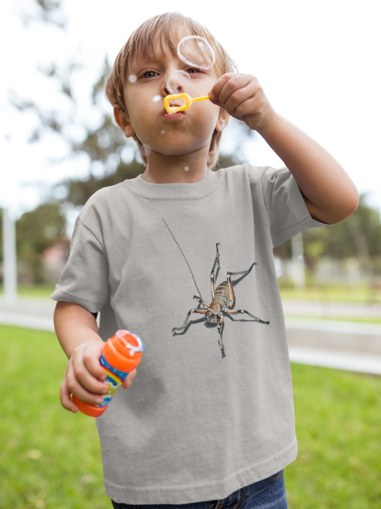A small child wearing a grey marle t-shirt with an all-over weta print is seen blowing bubbles, capturing a moment of childhood fun.