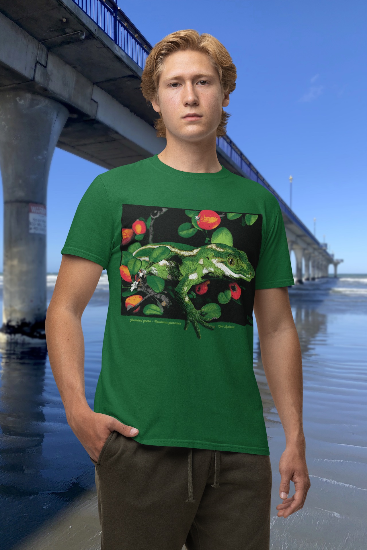 A man wearing a forest green Surface Active jewelled gecko t-shirt beside New Brighton Pier.