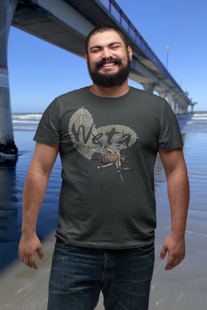 A man smiling beside New Brighton Pier, wearing a black marle t-shirt with a detailed Giant Weta print.