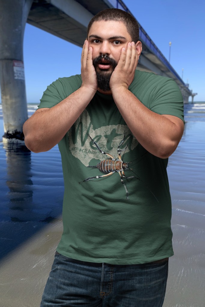 A man clutches his face in faux surprise wearing his forest green giant weta t-shirt beside New Brighton Pier.
