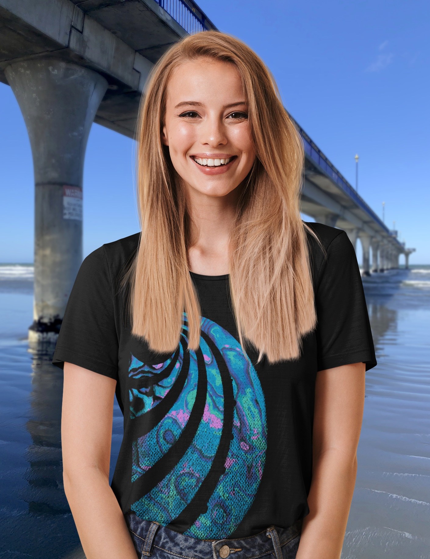 A smiling young woman beside New Brighton Pier wearing a black Surface Active t-shirt with a colourful Paua shell design.