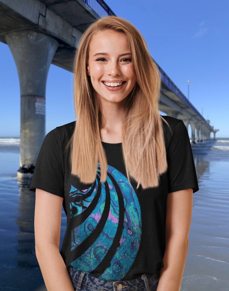 A smiling young woman beside New Brighton Pier wearing a black Surface Active t-shirt with a colourful Paua shell design.
