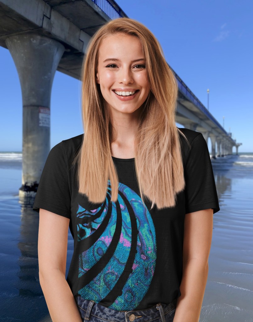 A smiling young woman beside New Brighton Pier wearing a black Surface Active t-shirt with a colourful Paua shell design.