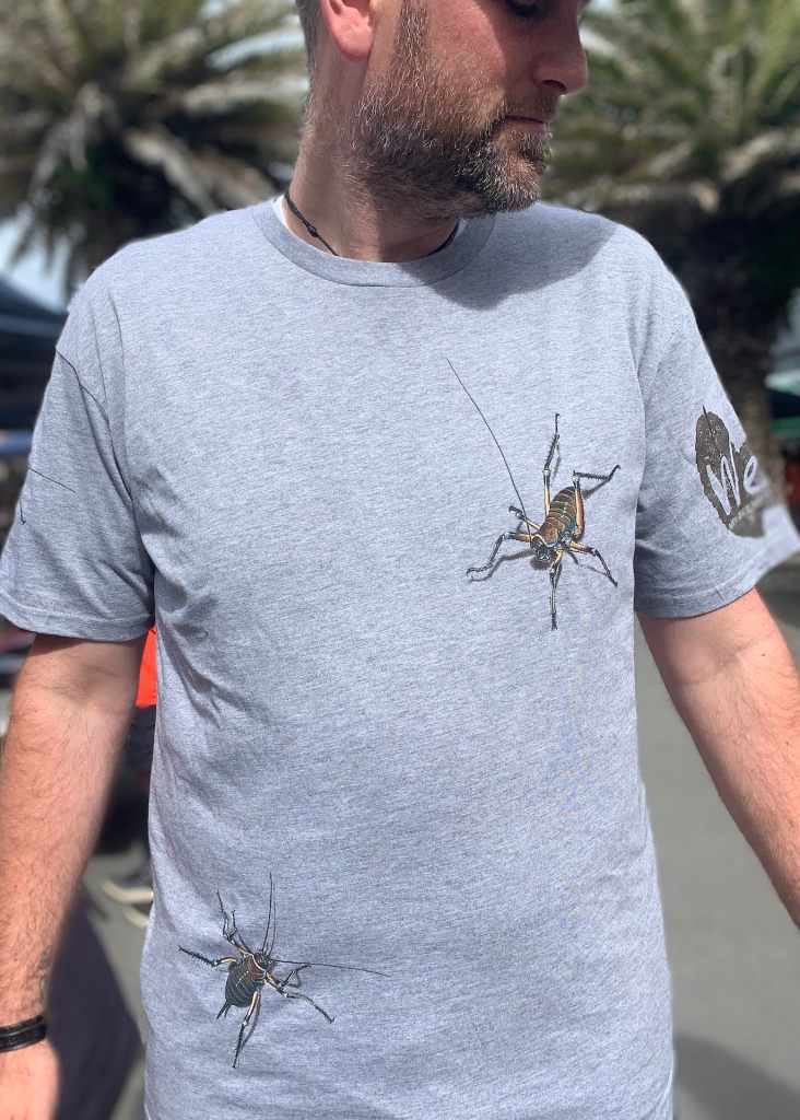 Ben, a regular customer at the New Brighton Seaside Market, wearing a grey marle t-shirt with an all-over Bluff weta print, showcasing Shaun Waugh’s Surface Active New Zealand Nature T-shirt stall in Brighton Mall on Saturday mornings.